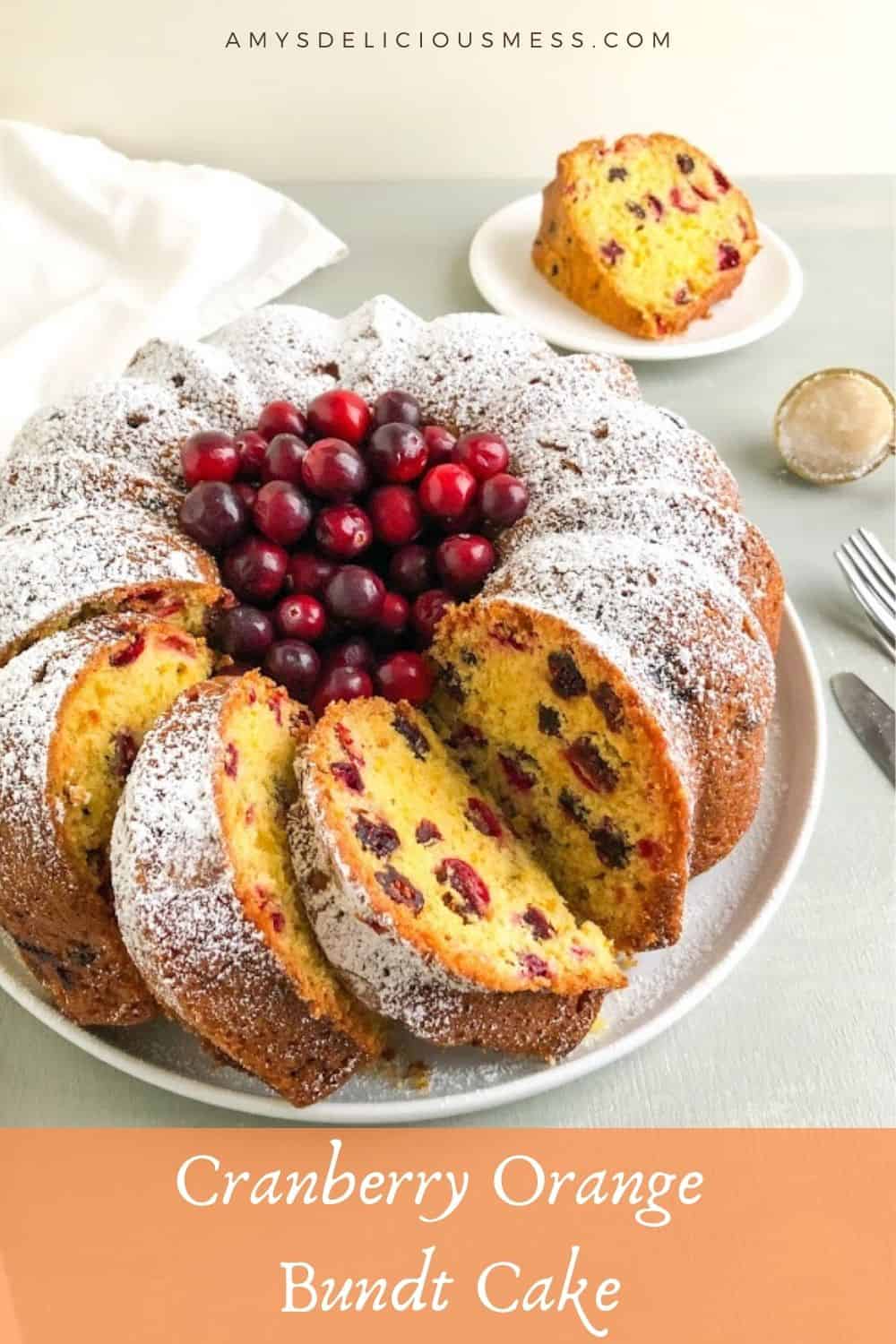 Whole bundt cake with a few slices dusted with powdered sugar on large round white plate with whole fresh cranberries in the center, next to white kitchen towel. One slice of cake on small round white plate and gold tea strainer with powdered sugar in the background.