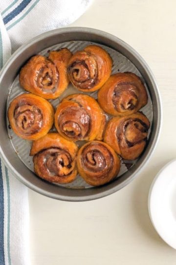 crescent "cinnamon rolls" in round metal cake pan next to white kitchen towel with dark and light blue stripes and small round white plates with silver cocktail forks