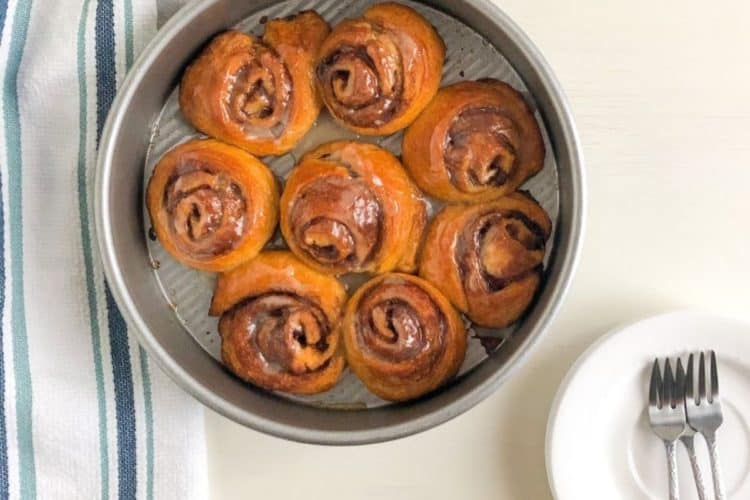 crescent "cinnamon rolls" in round metal cake pan next to white kitchen towel with dark and light blue stripes and small round white plates with silver cocktail forks