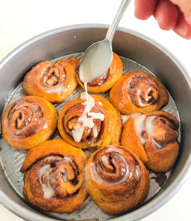 round metal cake pan of cinnamon rolls, hand holding small silver spoon drizzling icing