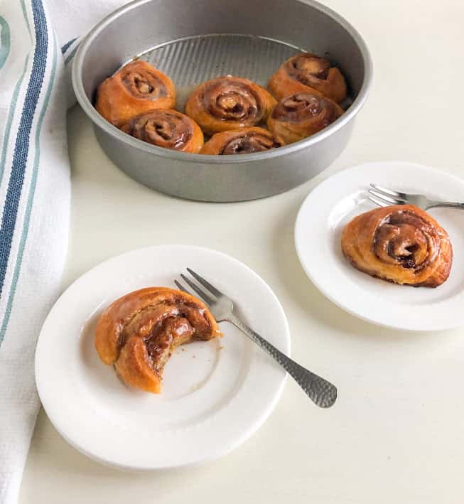 two round white plates with cinnamon rolls and cocktail fork with one cinnamon roll missing a bite. Round silver cake pan with remaining cinnamon rolls in the background next to kitchen towel with light and dark blue stripes