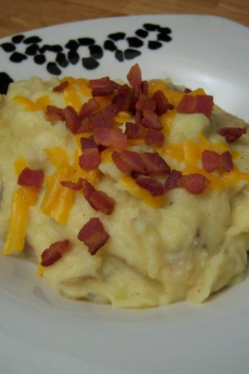Loaded mashed potatoes topped with shredded cheddar cheese and bacon bits in square bowl with black flowers