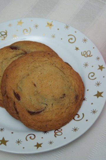 The Famous NY Times Chocolate Chip Cookies on small round plate with gold stars and swirls on striped cream place mat with plastic pink cup filled with milk