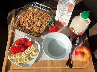 Baked apple oatmeal in square glass baking dish on wooden board with milk container, maple syrup, bowls, spoons, apple, sliced strawberries and banana on square plate