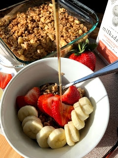 Baked apple oatmeal in square glass dish, white bowl with baked apple oatmeal topped with sliced banana and sliced strawberries.  Maple syrup drizzling on bowl of oatmeal