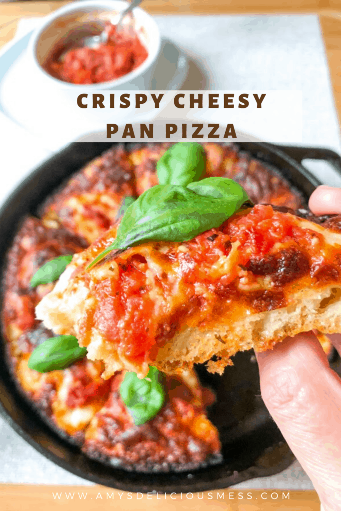 Crispy cheesy pan pizza topped with basil leaves in round cast iron pan on wooden board with small white round ramekin of pizza sauce on small square plate holding one slice of pizza