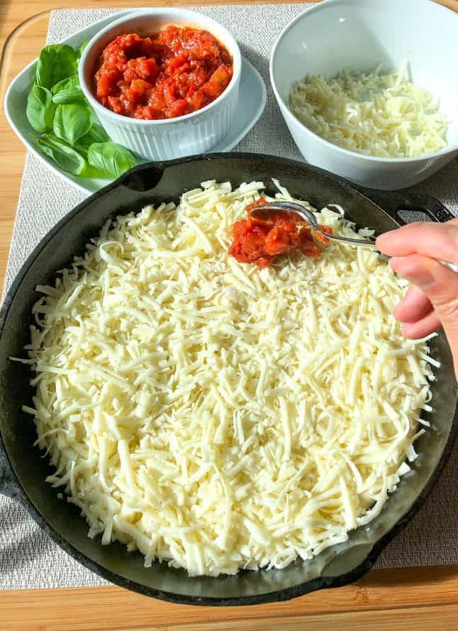 Pizza dough topped with mozzarella in round cast iron pan on wooden board with medium round white bowl with shredded mozzarella cheese, small round white ramekin with pizza sauce and spoon on small square plate with fresh basil leaves