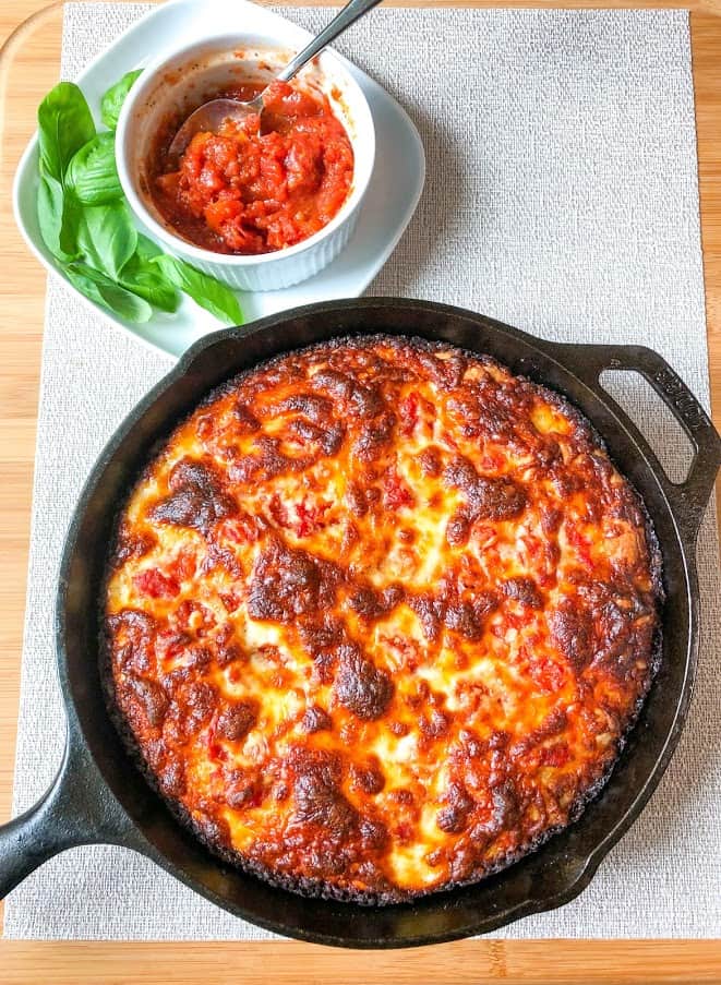 Baked cheesy crispy pan pizza in round cast iron pan on off white placement on wooden cutting board with small ramekin of pizza sauce with spoon on square plate with fresh basil leaves