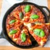Crispy cheesy pan pizza topped with basil leaves in round cast iron pan on wooden board