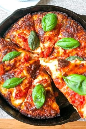 Crispy cheesy pan pizza topped with basil leaves in round cast iron pan on wooden board