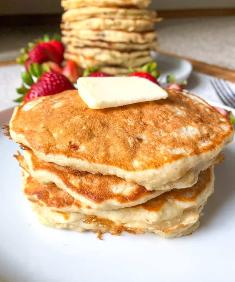 Banana oatmeal pancakes with pat of butter on round white plate with whole and quartered strawberries.  Fork and stack of banana oatmeal pancakes with whole and quartered strawberries in background