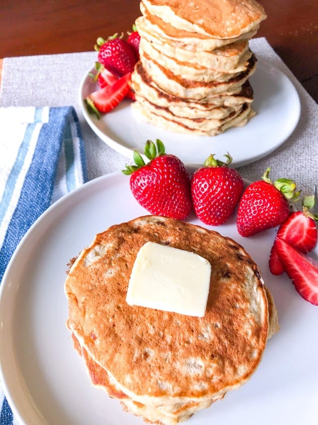 Banana oatmeal pancakes with pat of butter on round white plate with whole and quartered strawberries.  Blue and white striped towel and stack of banana oatmeal pancakes with whole and quartered strawberries in background.