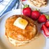 Stack of banana oatmeal pancakes on round white plate with pat of butter, cut strawberries, maple syrup drizzle on top of pancakes, stack of pancakes in background with strawberries
