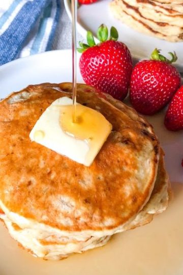 Stack of banana oatmeal pancakes on round white plate with pat of butter, cut strawberries, maple syrup drizzle on top of pancakes, stack of pancakes in background with strawberries