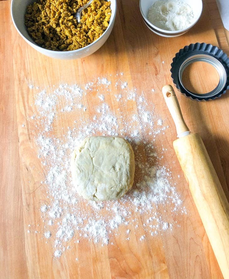 Turkey curry filling in a round gray bowl with spoon, flour in a small round stripe bowl, dough on wooden board dusted with flour next to wooden rolling pin
