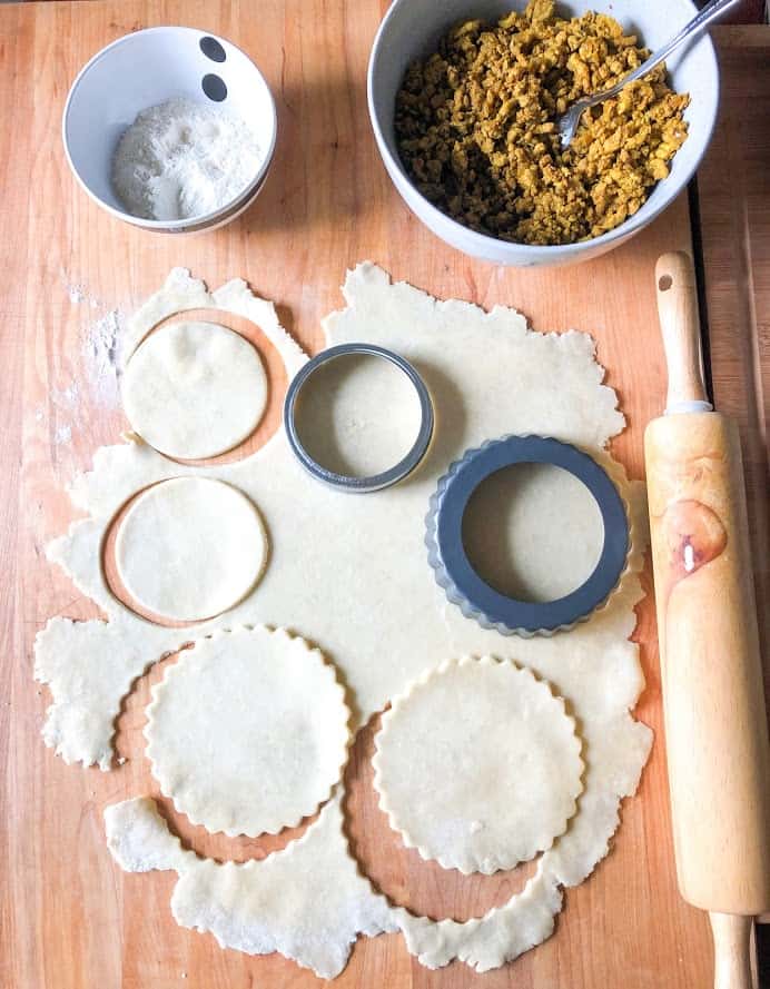 Turkey curry filling in a round gray bowl with spoon, flour in a small round stripe bowl, rolled dough on wooden board dusted with flour next to wooden rolling pin. Circles cut out with mason jar lid and mini tart pan