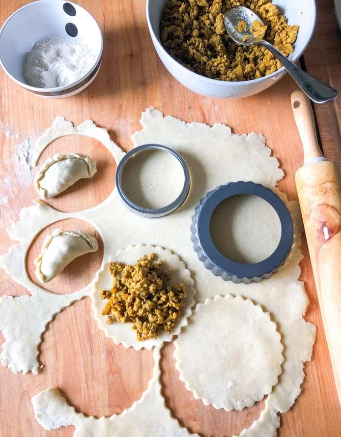 Turkey curry filling in a round gray bowl with spoon, flour in a small round stripe bowl, rolled dough on wooden board dusted with flour next to wooden rolling pin. Circles cut out with mason jar lid and mini tart pan. Formed empanadas laying on dough.
