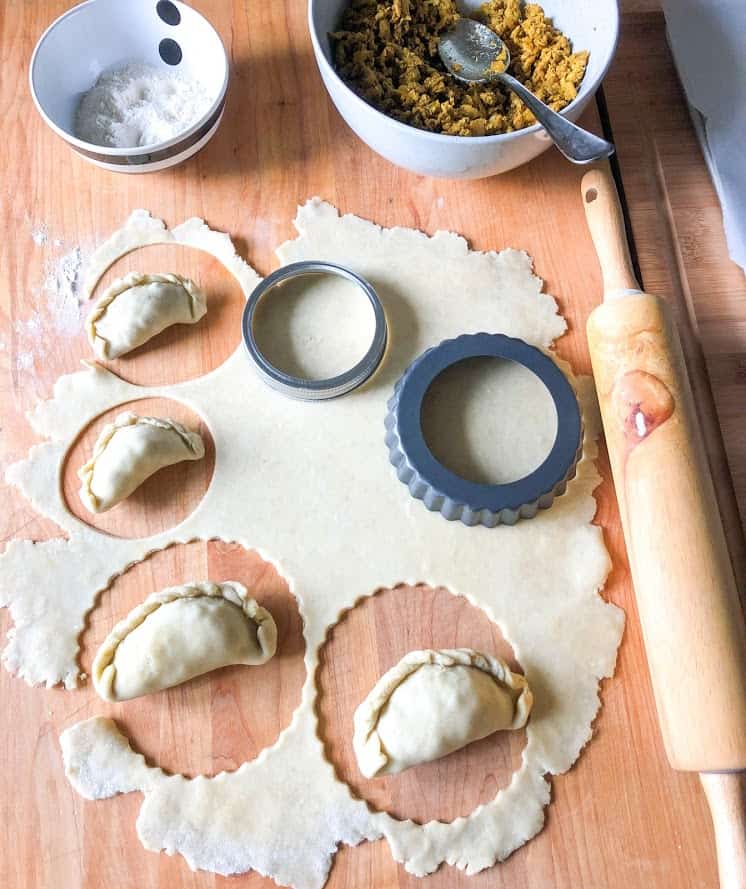 Turkey curry filling in a round gray bowl with spoon, flour in a small round stripe bowl, rolled dough on wooden board dusted with flour next to wooden rolling pin. Circles cut out with mason jar lid and mini tart pan. Formed empanadas placed in circle cutouts in dough