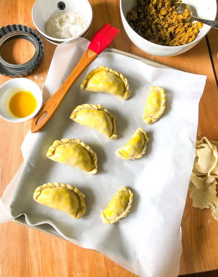 Turkey curry filling in a round gray bowl with spoon, flour in a small round stripe bowl, Assembled empanadas on parchment lined sheet tray with egg wash in small white round bowl.  Red pastry brush laying on sheet pan with egg washed empanadas.  Dough scraps on cutting board