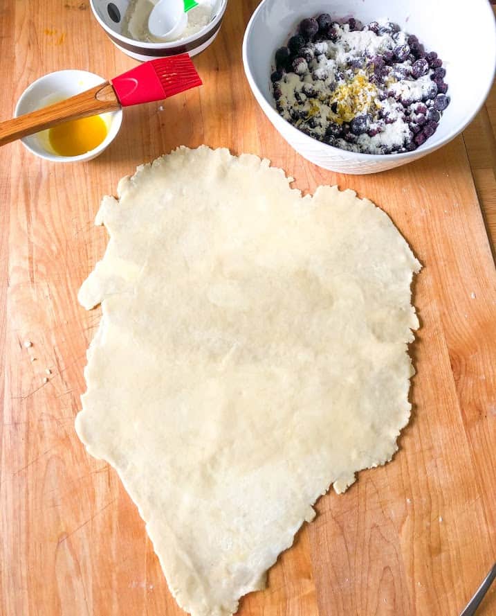 easy lemon blueberry galette, dough rolled out on wooden cutting board, egg wash in small round bowl, red pastry brush, round white bowl with frozen blueberry galette filling