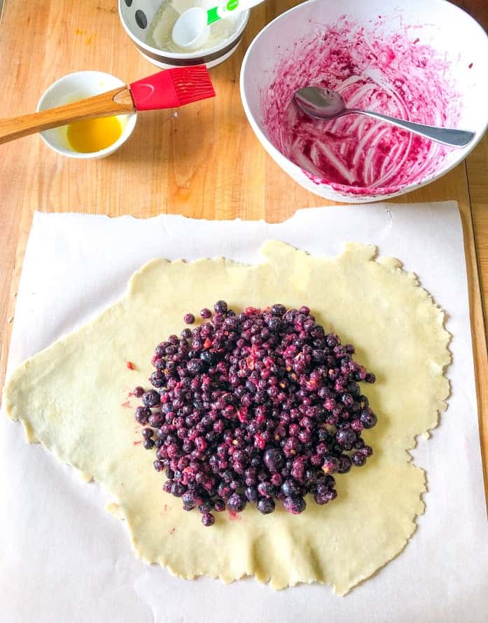 easiest blueberry lemon galette, dough rolled out on wooden cutting board, topped with frozen blueberry galette filling, egg wash in small round bowl, red pastry brush, empty round white bowl with frozen blueberry juices