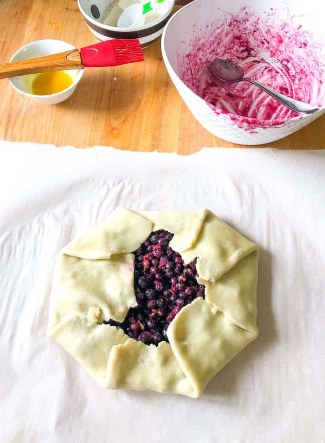 easiest blueberry lemon galette, rolled out dough on wooden cutting board, egg wash in small round bowl, red pastry brush, round white bowl with frozen blueberry galette filling