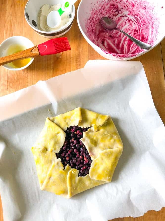 easiest blueberry lemon galette, galette formed on parchment paper on baking sheet on wooden cutting board, topped with frozen blueberry galette filling, egg washed amd sprinkled with sugar in small round bowl, red pastry brush, empty round white bowl with frozen blueberry juices