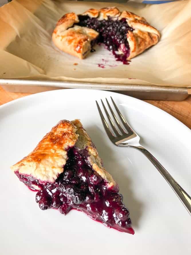 blueberry lemon galette slice on round white plate with fork, whole blueberry galette on parchment paper lined baking sheet in background