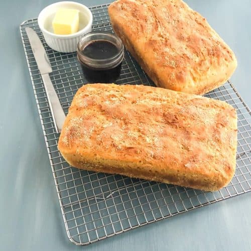 Two loaves of English muffin bread on a metal cooling rack with jar of jam and small ramekin of butter with silver butter knife