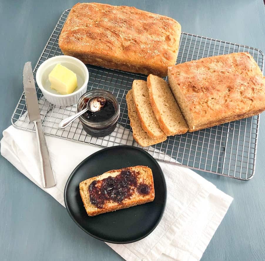 English muffin loaves on silver metal cooling rack, one loaf sliced, small ramekin of butter, small jar of jam with small silver spoon on top, silver butter knife, white kitchen towel, small round black plate with slice of English muffin bread with butter and jam
