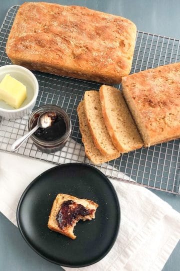 English muffin loaves on silver metal cooling rack, one loaf sliced, small ramekin of butter, small jar of jam with small silver spoon on top, silver butter knife, white kitchen towel, small round black plate with slice of English muffin bread with butter and jam, half eaten