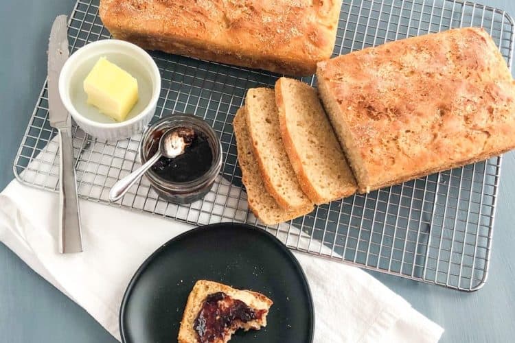 English muffin loaves on silver metal cooling rack, one loaf sliced, small ramekin of butter, small jar of jam with small silver spoon on top, silver butter knife, white kitchen towel, small round black plate with slice of English muffin bread with butter and jam, half eaten