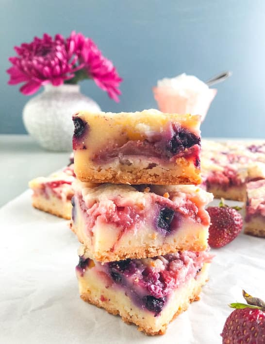 Mixed berry lemon pie bars stacked on parchment paper, with whole strawberries and more mixed berry lemon pie bars in the back ground, vintage pink depression glass ice cream dish with whipped cream, small white vase with gray cracks and purple China aster flowers