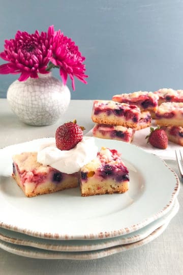 Mixed berry lemon pie bar squares on light blue plate topped with whipped cream and whole strawberry, with stacks of mixed berry lemon pie bars in the back ground on parchment paper with whole strawberries, small vase with gray cracks and purple china aster, stack of silver dinner forks