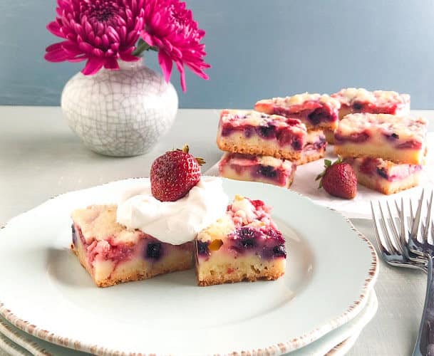 Mixed berry lemon pie bar squares on light blue plate topped with whipped cream and whole strawberry, with stacks of mixed berry lemon pie bars in the back ground on parchment paper with whole strawberries, small vase with gray cracks and purple china aster, stack of silver dinner forks