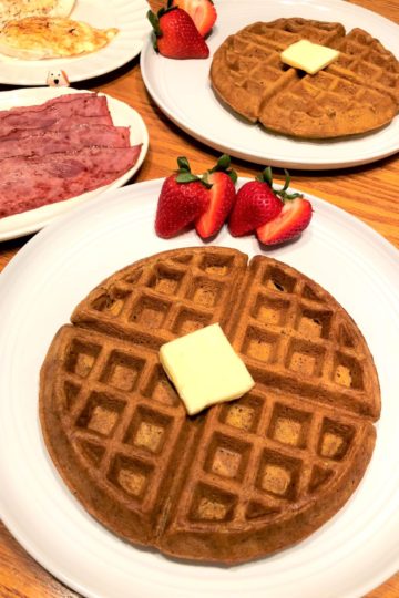 Pumpkin waffle on white plate with pat of butter and cut strawberries. Eggs and turkey bacon on plates in the background