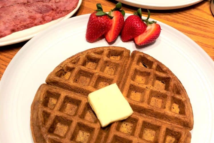 Pumpkin waffle on white plate with pat of butter and cut strawberries. Eggs and turkey bacon on plates in the background