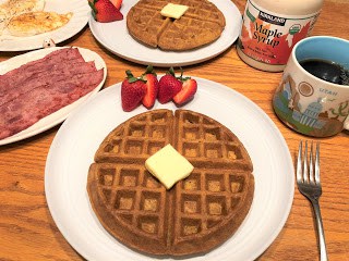 Pumpkin waffle on round white plate with pat of butter and cut strawberries, Turkey bacon and eggs on plates in the background.  Cup of coffee and maple syrup bottle in the background
