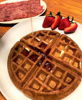 Pumpkin waffle on round white plate with cut strawberries, drizzling syrup with turkey bacon on plate in background