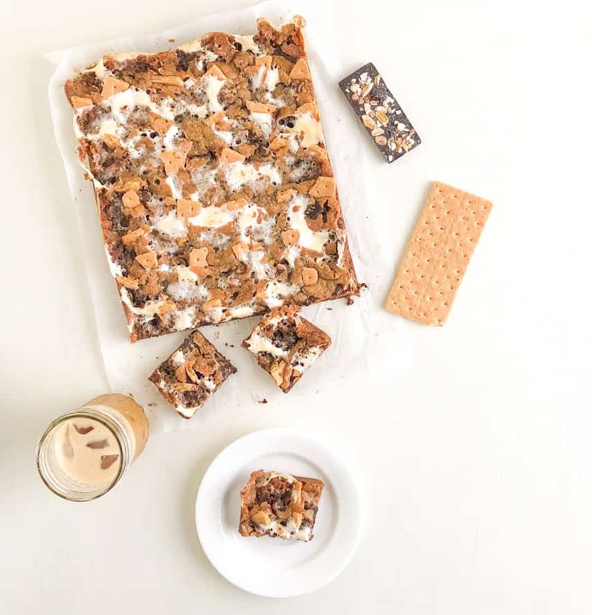 Gooey browned butter s'mores bar on small white round plate with since graham cracker sheet, ice latte in mason jar, chocolate bar with nuts, two s'mores bars and sheet of s'mores bars on parchment paper