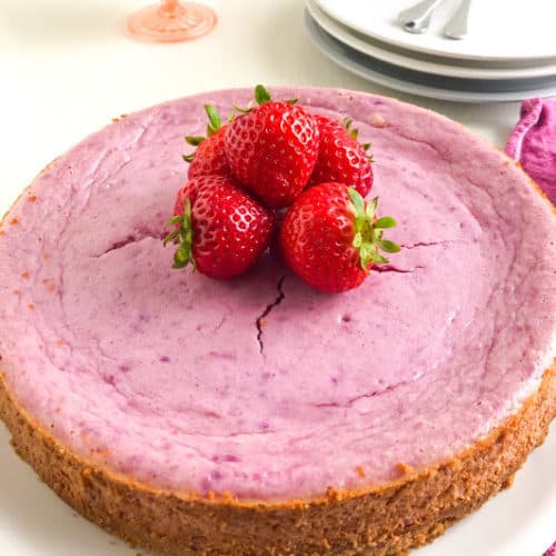 Purple sweet potato cheesecake on round white place topped with whole strawberries on pink kitchen towel, with two round white plates and one round gray plate stacked in background with small silver forks on top, pink vintage dessert glass with whipped cream in background