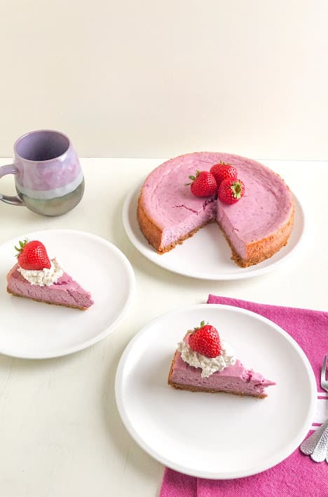 Two slices of purple sweet potato cheesecake on round white plates topped with whipped cream and whole strawberries on a pink kitchen towel with silver cocktail forks, purple and gray coffee mug and remaining cheesecake on large round white plate topped with whole strawberries in the background