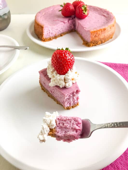 Slice of purple sweet potato cheesecake on a round white plate topped with whipped cream and a whole strawberry, bite of cheesecake with whipped cream on a small sliver cocktail fork on a pink kitchen towel, remaining cheesecake on a large round white plate topped with strawberries, purple and gray coffee mug and white plate with silver fork handle in the background