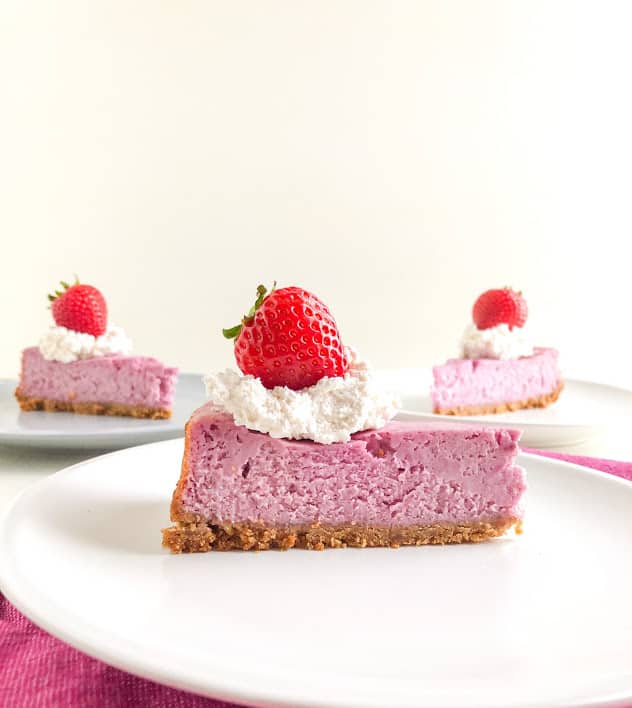 Purple sweet potato cheesecake slice topped with whipped cream and a whole strawberry on round white plate with two cheesecake slices in the background on a round gray plate and a round white plate both topped with whipped cream and a whole strawberry