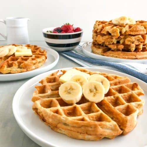Simple banana oatmeal waffle on small white round plate with pat of butter, sliced strawberries, powdered sugar, small pitcher pouring maple syrup on waffles, small round white bowl with black stripes with mixed berries, stack of waffle on small white round plate in background