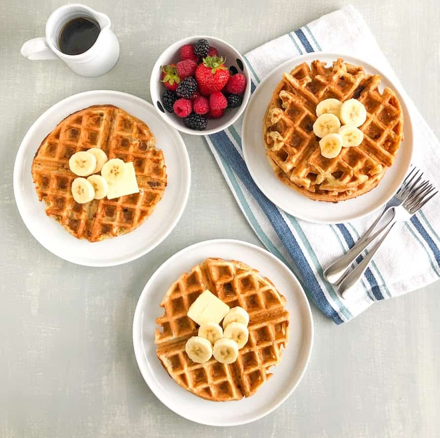 Simple banana oatmeal waffles on small white round plates with pat of butter, sliced bananas, small white pitcher of maple syrup, small round white bowl with mixed berries, stack of waffle on small white round plate on white and blue stripped kitchen towel with silver forks