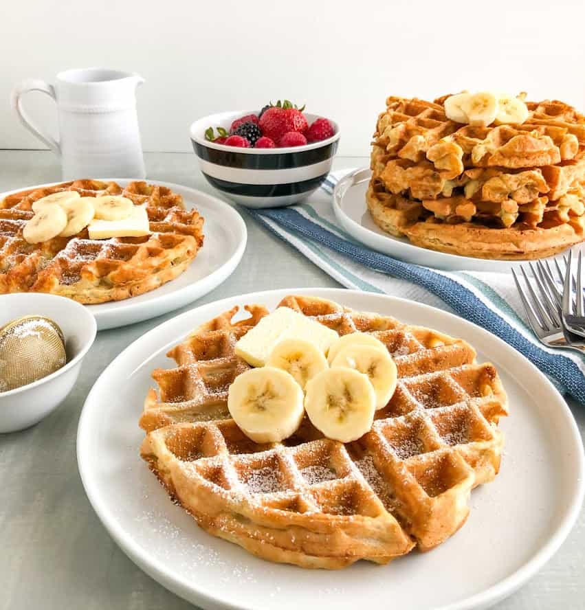 Simple banana oatmeal waffles on small white round plates with pat of butter, sliced bananas, small white pitcher of maple syrup in background, small round white bowl with black stripes with mixed berries, stack of waffle on small white round plate in background on white and blue stripped kitchen towel with silver forks