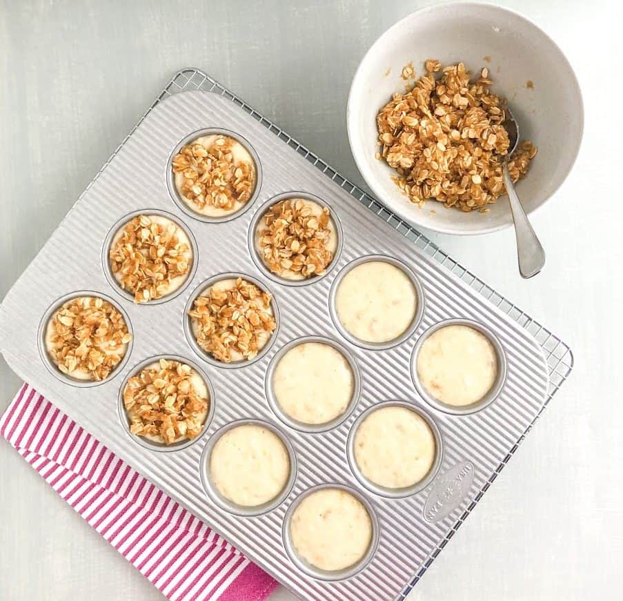Banana muffin batter in silver muffin pan on silver wire cooling rack on pink striped kitchen towel, medium gray bowl of oatmeal topping with silver spoon on the side
