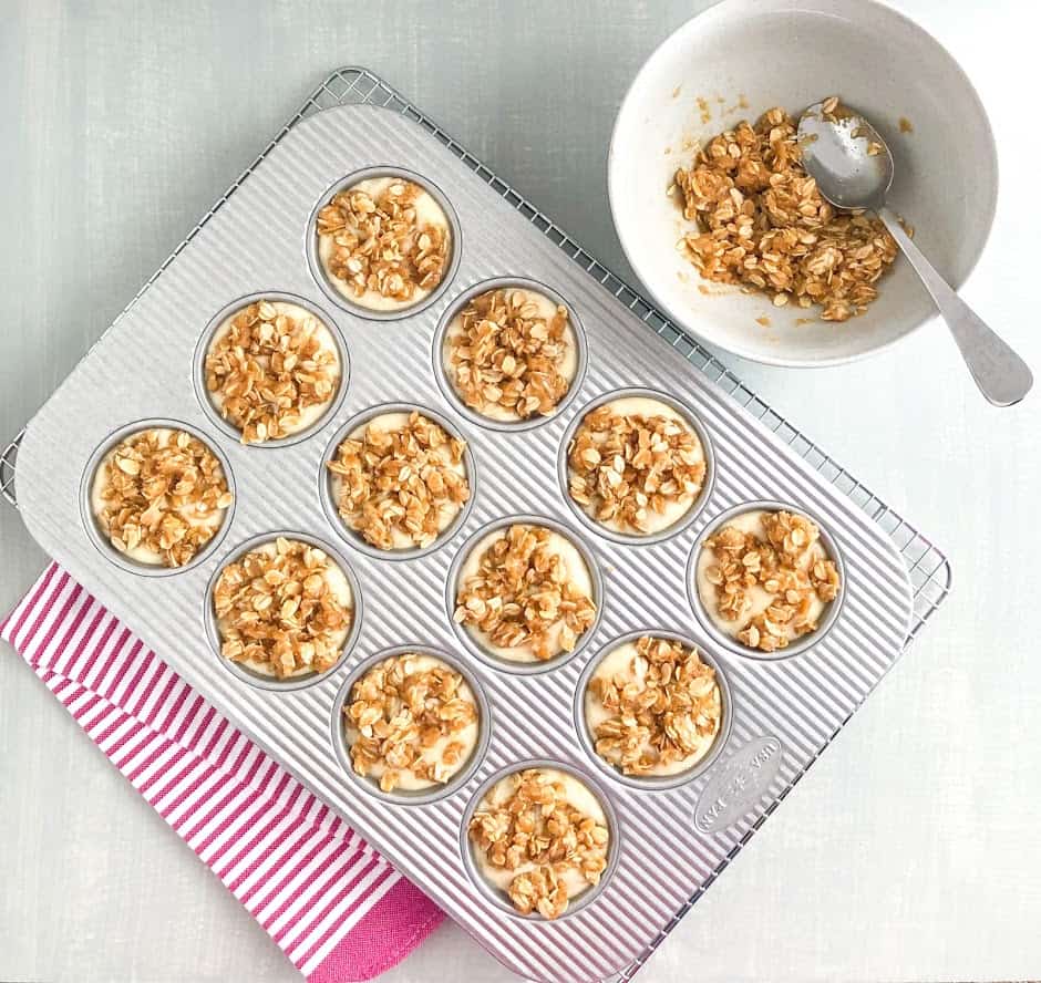Banana muffin batter with oatmeal topping in silver muffin pan on silver wire cooling rack on pink striped kitchen towel, medium gray bowl of oatmeal topping with silver spoon on the side