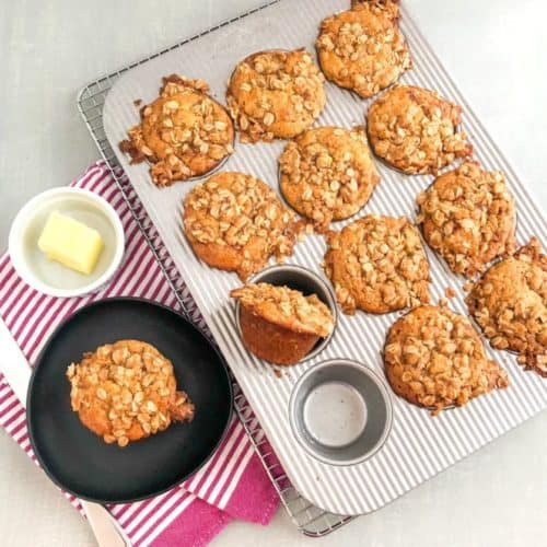 Banana muffin with oatmeal topping on small black plate on a pink striped kitchen towel next to a small round white ramekin with butter, silver butter knife, muffin pan of banana muffins on silver cooling rack